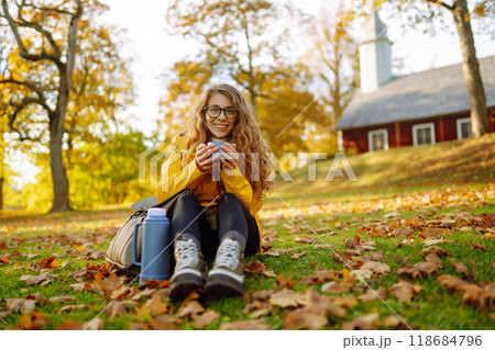 Woman traveler in yellow coat sits in clearing among yellow leaves and drinks hot drink from thermos Woman traveler in yellow coat sits in clearing among yellow leaves and drinks hot drink from thermos 118684796