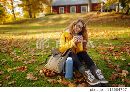 Woman traveler in yellow coat sits in clearing among yellow leaves and drinks hot drink from thermos Woman traveler in yellow coat sits in clearing among yellow leaves and drinks hot drink from thermos 118684797
