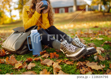 Woman traveler in yellow coat sits in clearing among yellow leaves and drinks hot drink from thermos Woman traveler in yellow coat sits in clearing among yellow leaves and drinks hot drink from thermos 118684798