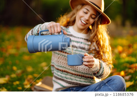 Curly woman with thermos in clearing among fallen leaves in autumn park, enjoying hot drink. 118685376