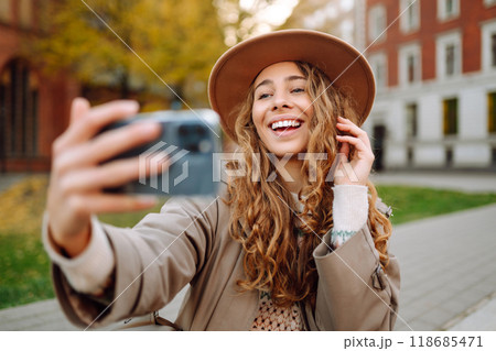 Stylish woman in a raincoat and hat takes a selfie outdoors in autumn on the streets of the city. Stylish woman in a raincoat and hat takes a selfie outdoors in autumn on the streets of the city. 118685471