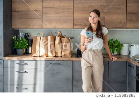 Young woman with smart phone in hand next to paper bags full of food in kitchen. Groceries ordered online and delivered by courier Young woman with smart phone in hand next to paper bags full of food in kitchen. Groceries ordered online and delivered by courier 118688865
