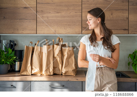 Young woman with receipt from the store next to paper bags full of food in kitchen. Groceries ordered online and delivered by courier 118688866