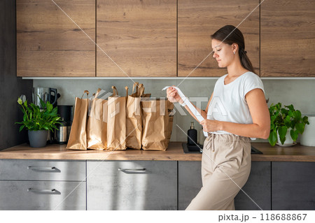 Young woman with receipt from the store next to paper bags full of food in kitchen. Groceries ordered online and delivered by courier 118688867