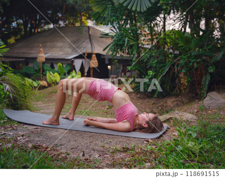 Beautiful woman practices yoga by pool in morning in Thailand. Young female in sportswear in hotel exotic garden. perfect flexible body recreating doing yoga poses feeling inner peace and vitality 118691515