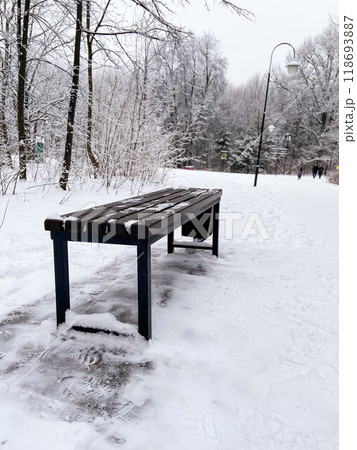 Solitary bench stands in a snow covered park during winter, inviting quiet reflection and peace. Winter landscape captures the beauty of cold season in nature Solitary bench stands in a snow covered park during winter, inviting quiet reflection and peace. Winter landscape captures the beauty of cold season in nature 118693887
