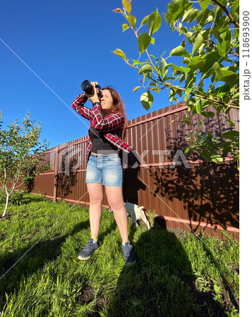 Millennial woman taking photo in a sunny backyard with a small tree and pet dog. Bright and clear blue sky providing a perfect backdrop for outdoor photography. Photographer, connection with nature 118693900