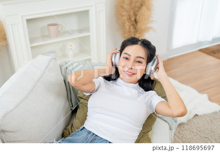 Beautiful Asian woman lie on sofa in living room of her house enjoy to listen song with earphone and smile at camera. Beautiful Asian woman lie on sofa in living room of her house enjoy to listen song with earphone and smile at camera. 118695697
