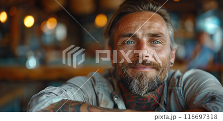 Smiling middle-aged man with tattoos and a  beard leaning forward in a warm, softly lit indoor setting 118697318