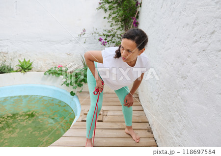 Woman exercising outdoors by a small round pool on a wooden deck, wearing white and mint activewear, focused on fitness routine Woman exercising outdoors by a small round pool on a wooden deck, wearing white and mint activewear, focused on fitness routine 118697584