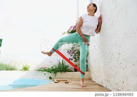 Woman exercising with resistance band outdoors in a sunny garden, performing side leg lifts for fitness and flexibility 118698485