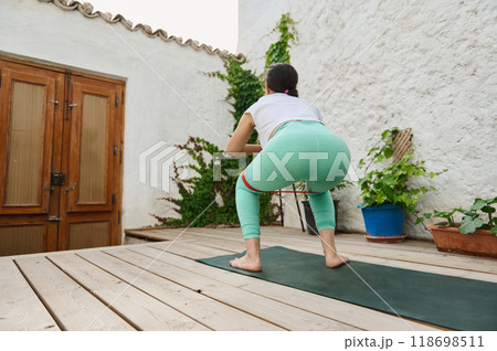 Woman exercising with resistance band on outdoor wooden deck, focusing on fitness and strength training in a serene garden setting 118698511