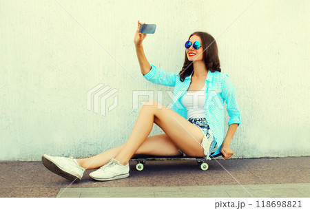 Modern happy smiling young woman taking selfie by smartphone sitting on skateboard on city street Modern happy smiling young woman taking selfie by smartphone sitting on skateboard on city street 118698821