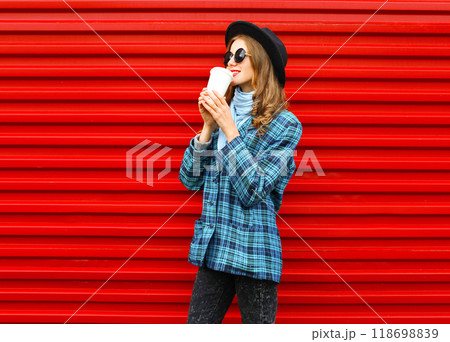 Fashion pretty woman drinks coffee from cup in a black hat coat jacket over colorful red background Fashion pretty woman drinks coffee from cup in a black hat coat jacket over colorful red background 118698839