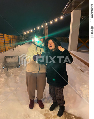 60 year old mother and her 30 year old daughter enjoy a winter night outside a cozy house and celebrating New Year holding a sparkler amidst snow and festive string lights. Heartwarming scene of 118699849