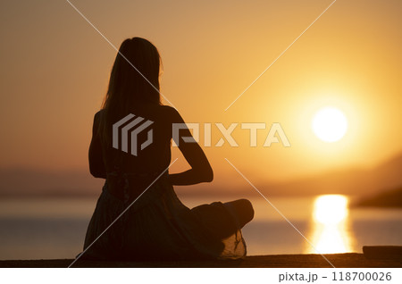 Young woman in summer dress meditating at beautiful warm colored sunrise by the sea 118700026