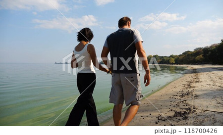 Couple holding hands of each other and walking at coast. Happy pair going along seashore at sunny day. Lovers spending time together at resort. Concept of vacation or holiday. Slow mo 118700426