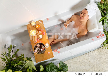 A young beautiful woman lies in a bath with foam and rose petals, reads a book and drinks champagne. 118703029