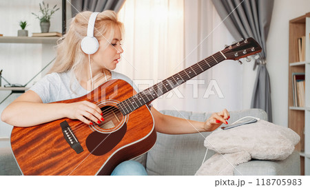 Guitar playing. Professional woman. Home lesson. Inspired lady in headphones tuning strings of musical instrument using mobile phone light room interior. 118705983