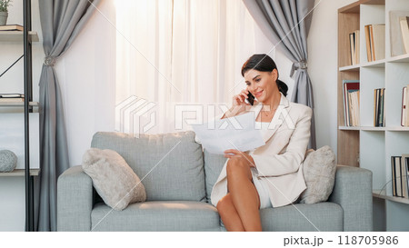Distance agreement. Female chief. Mobile chatting. Positive elegant woman talking smartphone holding paper documents sitting sofa light room interior. 118705986