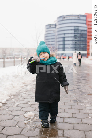 Boy child in a blue cap and a black coat is eating candy on a snowy sidewalk 118706845