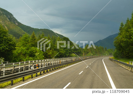 Winding road through lush green mountains under dramatic stormy clouds in early afternoon light 118709045