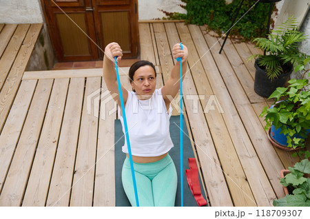 Woman practicing yoga with resistance band on wooden deck, focusing on strength and flexibility in a serene outdoor setting 118709307