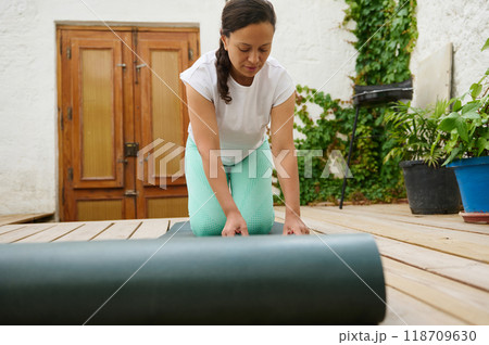 Woman setting up yoga mat on wooden patio for outdoor exercise in a peaceful garden setting Woman setting up yoga mat on wooden patio for outdoor exercise in a peaceful garden setting 118709630