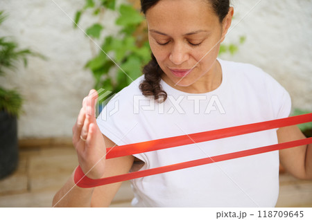 Focused woman exercising with red resistance band outdoors, promoting fitness, strength, and healthy lifestyle in a serene garden setting Focused woman exercising with red resistance band outdoors, promoting fitness, strength, and healthy lifestyle in a serene garden setting 118709645