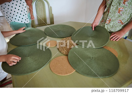Family bonding over setting a dining table with green placemats and woven decor for a casual gathering at home Family bonding over setting a dining table with green placemats and woven decor for a casual gathering at home 118709917
