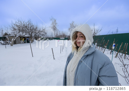 Young woman enjoying snowy garden on winter day, smiling warmly amidst frosty embrace of nature. Standing in backyard of her house cute woman smiles looking at camera. Copy space. 118712865