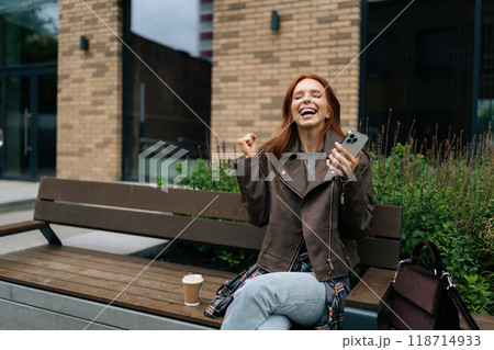 Cheerful redhead woman receiving unexpected good news in social media app using smartphone sitting on urban bench on summer day. Happy female celebrating success with joy and positive vibes. Cheerful redhead woman receiving unexpected good news in social media app using smartphone sitting on urban bench on summer day. Happy female celebrating success with joy and positive vibes. 118714933