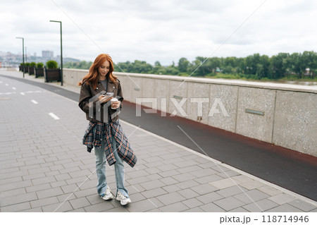 Remote view of pretty young woman happily strolling by riverfront, enjoying coffee break while checking phone on cloudy day. Cheerful redhead female in leather jacket enjoying free time outdoors. Remote view of pretty young woman happily strolling by riverfront, enjoying coffee break while checking phone on cloudy day. Cheerful redhead female in leather jacket enjoying free time outdoors. 118714946