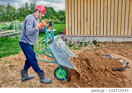 Construction worker fills area around septic tank manhole cover with sand using wheelbarrow. 118715543