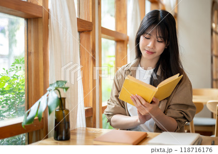 A charming Asian woman sits in a minimalist coffee shop, enjoying her reading in the cafe. A charming Asian woman sits in a minimalist coffee shop, enjoying her reading in the cafe. 118716176