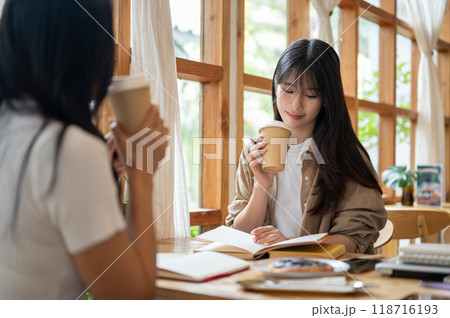 Two cheerful Asian college students are enjoying coffee while reading together in a coffee shop. Two cheerful Asian college students are enjoying coffee while reading together in a coffee shop. 118716193