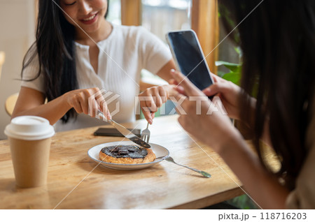A close-up of a girl taking a picture of a dessert with her smartphone while her friend is eating. 118716203