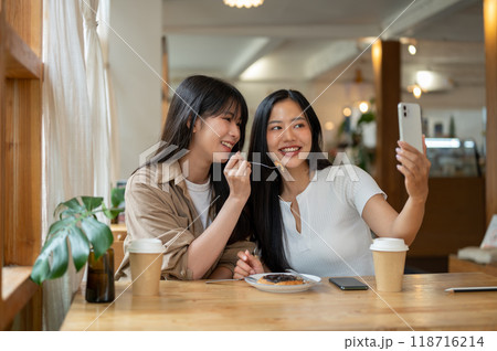 Two beautiful Asian women are enjoying taking pictures together while hanging out at a coffee shop. 118716214