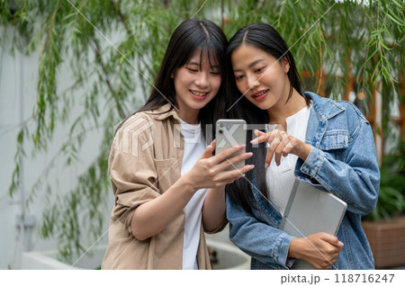 Two cheerful Asian female friends are watching something on a smartphone and enjoying a conversation Two cheerful Asian female friends are watching something on a smartphone and enjoying a conversation 118716247