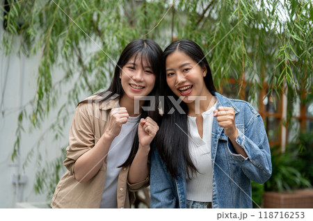 Two beautiful Asian women standing in a green garden, showing their fist and smiling at the camera. Two beautiful Asian women standing in a green garden, showing their fist and smiling at the camera. 118716253