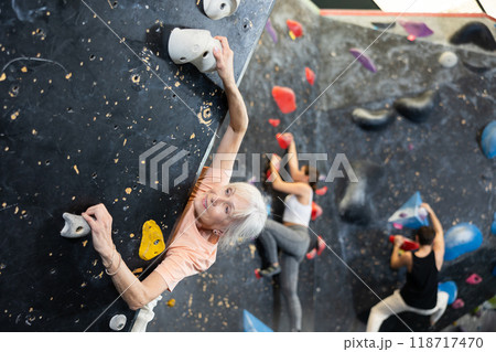 Positive focused mature woman exercising on black climbing wall without safety belts and preparing for summer mountain hiking in game center indoors Positive focused mature woman exercising on black climbing wall without safety belts and preparing for summer mountain hiking in game center indoors 118717470