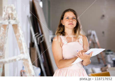 Portrait of positive woman next to a stepladder with documents in her hands checks the quality of the completed repairs in cottage room 118717652