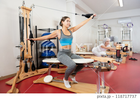 Sports club girl client on reformer with cable and rope performs exercise to stretch muscles of arms 118717737