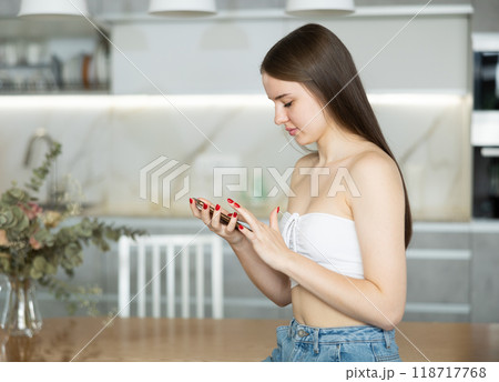 Young teen girl sat down on edge of empty dining table with smartphone in hands. Young teen girl sat down on edge of empty dining table with smartphone in hands. 118717768