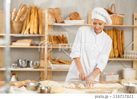 Woman works in bakery as baker, cuts dough into portions, forms pieces of dough to create buns. 118717821