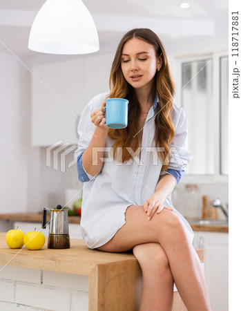 Woman sitting on countertop at kitchen and looking in cup Woman sitting on countertop at kitchen and looking in cup 118717881