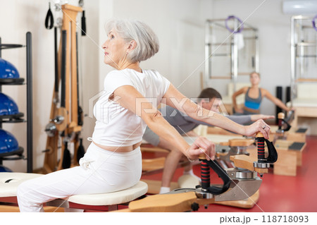 Elderly woman stretches her arms using pilates machine while sitting backwards 118718093
