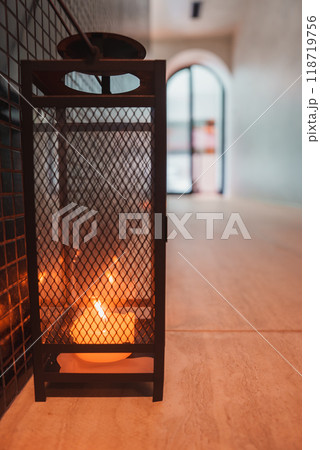 Close-up of a lit candle inside a black metal lantern with mesh design on a tiled floor. The blurred hallway and arched doorway suggest a high-end interior setting. Close-up of a lit candle inside a black metal lantern with mesh design on a tiled floor. The blurred hallway and arched doorway suggest a high-end interior setting. 118719756