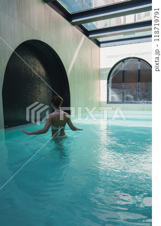 A woman in a white swimsuit wades through a serene, turquoise pool in a high-end hotel. The modern design features arched windows and a glass ceiling, creating an elegant ambiance. A woman in a white swimsuit wades through a serene, turquoise pool in a high-end hotel. The modern design features arched windows and a glass ceiling, creating an elegant ambiance. 118719791