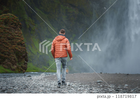 A solitary figure in an orange jacket walks towards the powerful Skogafoss waterfall in Iceland, surrounded by lush green cliffs and misty atmosphere. A solitary figure in an orange jacket walks towards the powerful Skogafoss waterfall in Iceland, surrounded by lush green cliffs and misty atmosphere. 118719817